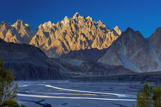 View of the majestic Passu Cones mountain range with golden sunlight illuminating the jagged peaks above a wide riverbed, Hunza Nagar, Gilgit Baltistan, Pakistan.