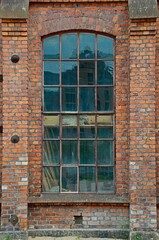 Obraz premium Weathered brick factory wall with tall arched industrial window, rusted metal frame, aged glass reflections, historic masonry texture, vertical architectural detail, urban heritage facade