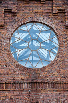 Large circular industrial window set into weathered red brick wall, featuring geometric glass panes and arched masonry details, creating strong symmetry, texture, heritage architecture appeal