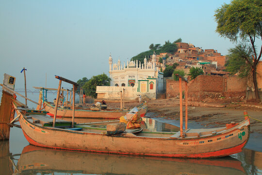View of traditional wooden boats moored on the banks of the Indus river with the historic white mosque and hillside houses of Kala Bagh town, Kalabagh, Punjab, Pakistan.