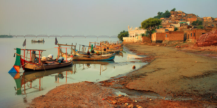 View of traditional wooden boats moored along the sandy banks of the Indus River with the historic town of Kalabagh and a mosque in the background, Kalabagh, Punjab, Pakistan.