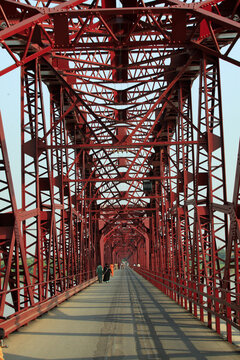 View of the historic red steel truss Kalabagh Railway Bridge from the colonial period spanning the Indus River with people walking along the path in Kalabagh, Punjab, Pakistan.