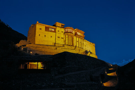 View of the illuminated Baltit Fort at night, featuring historic stone architecture set against a dark blue sky in Karimabad, Gilgit Baltistan, Pakistan.