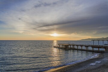 Fototapeta premium A peaceful golden sunset over the sea with a concrete pier and a sandy coastline featuring empty sunbeds