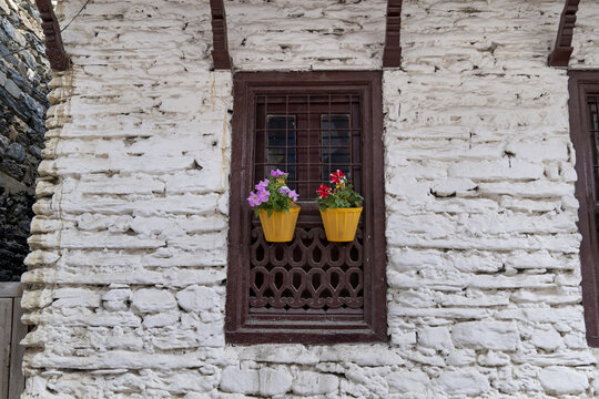 View of a traditional white stone wall with a dark wooden window frame featuring intricate lattice work and two yellow flower pots in Marpha, Jomsom, Gandaki Province, Nepal.