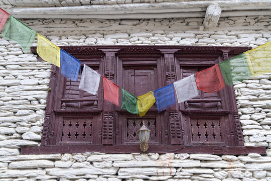 View of the traditional stone architecture and intricately carved wooden window frame of a building in Marpha decorated with colorful Tibetan prayer flags in Jomsom, Gandaki Province, Nepal.