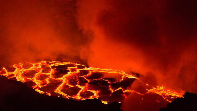 View of the glowing molten lava lake at Erta Ale volcano with cracks in the cooling crust and thick volcanic smoke rising into the night sky in Zone 2, Afar, Ethiopia.