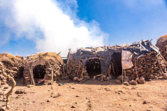 View of traditional stone huts with thatched and plastic roofs near the Erta Ale volcano, set against a clear blue sky with volcanic steam in Zone 2, Afar, Ethiopia.