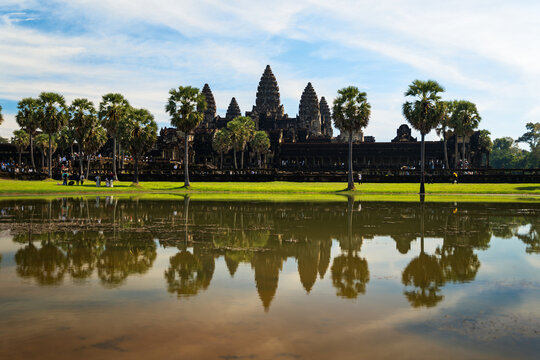 View of Angkor Wat temple complex with its reflection in the foreground pond, surrounded by palm trees under a blue sky with light clouds in Krong Siem Reap, Siem Reap Province, Cambodia.