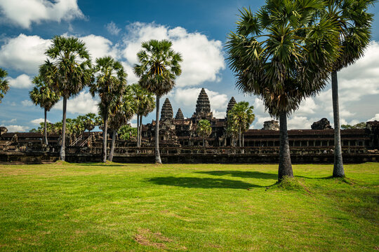 View of the ancient Angkor Wat temple complex surrounded by lush green lawns and tall palm trees under a bright blue sky in Krong Siem Reap, Siem Reap Province, Cambodia.