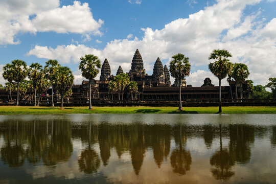 View of Angkor Wat temple reflected in the surrounding moat with palm trees under a blue sky with white clouds in Krong Siem Reap, Siem Reap Province, Cambodia.