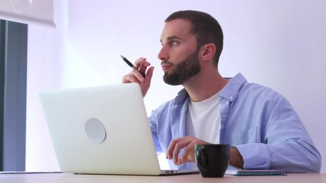 Young handsome freelancer thinking about a solution while working at his modern home office, looking for inspiration with a pensive expression on his face while using his laptop