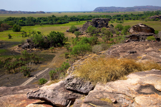 Two hikers traverse the rugged sandstone cliffs overlooking an expansive landscape of green plains and trees. They enjoy the scenic view in the quiet wilderness area.