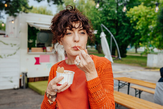 Playful middle-aged woman with braces tasting rolled ice cream near food truck