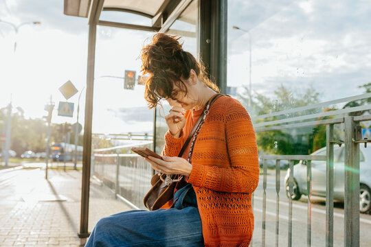 Thoughtful middle-aged woman using smartphone while waiting at city bus stop