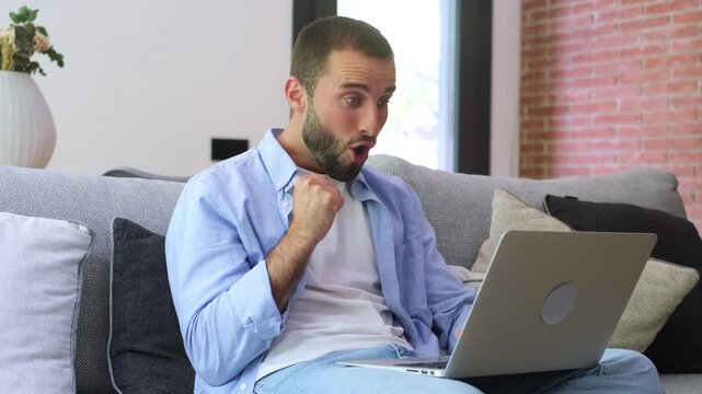 Overjoyed man sitting on the sofa celebrating a victory or business success while looking at his laptop, feeling excited and happy after receiving good news online in his modern apartment