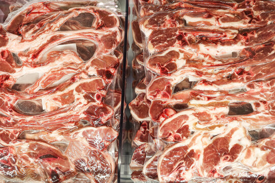 Raw lamb chops fill packages. Plastic wrap seals fresh meat. Butcher display forms a cold background. Marbled cuts reveal bone sections. Refrigerated food waits for retail customers.