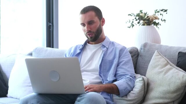 Handsome caucasian man relaxing on the sofa in his modern living room, working remotely from home and engaging in a business video conference call using his personal laptop