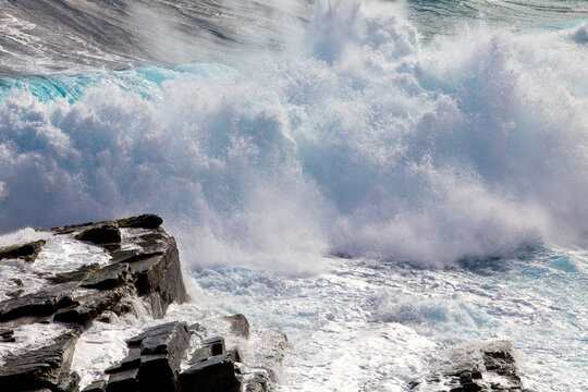 A powerful ocean wave crashes against the dark jagged rocks of the rugged coastline. The sea water churns into thick white foam as the tide surges with immense force.