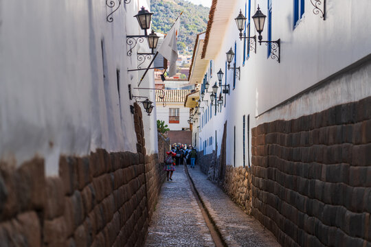 View of a narrow historic street with traditional Inca stone masonry walls, white colonial buildings, hanging lanterns, and a person in traditional clothing, Cusco, Cusco, Peru.