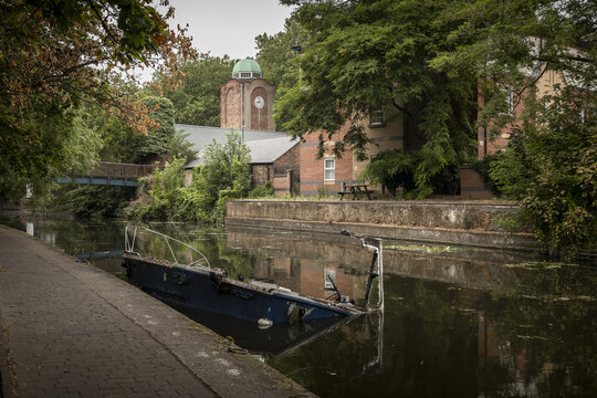 View of a partially submerged boat in the Nottingham canal, with historic brick buildings, a clock tower, and lush trees in Nottingham, England, United Kingdom.