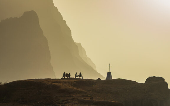 View of people sitting on a bench near a cross with dramatic cliffs in the background during golden hour in Ponta do Sol, Ribeira Grande, Cabo Verde.