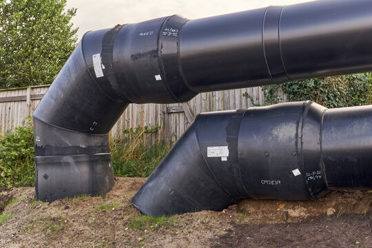 View of large black industrial pipes and elbow joints of the district heating system installed in the ground with a wooden fence in the background in Esbjerg, Denmark.
