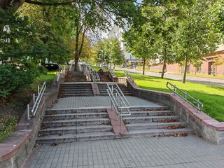 Obraz premium A staircase made of concrete tiles with metal railings has been built on the city street. There are buildings and trees nearby and the roadway is paved with paving stones. Sunny summer weather
