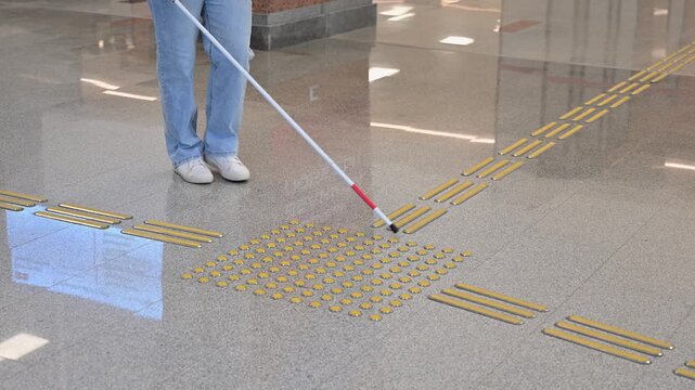 A blind woman walks on tactile paving. Close-up of her feet and a walking stick.