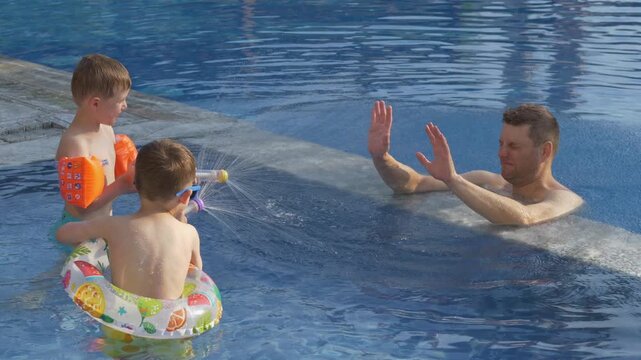 Father and two boys enjoy energetic water blaster game in sunny pool. Childhood adventure, outdoor recreation and joyful family holiday concept. 