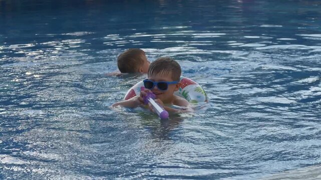 Young boy aims colorful water blaster and sprays water while playing in outdoor swimming pool. Imaginative play, active childhood, summer fun and joyful water games concept.