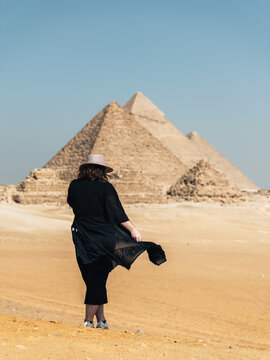 View of a person in a hat and black dress standing in the desert sand with the Giza Necropolis pyramids in the background under a clear blue sky in Al Haram, Giza Governorate, Egypt.