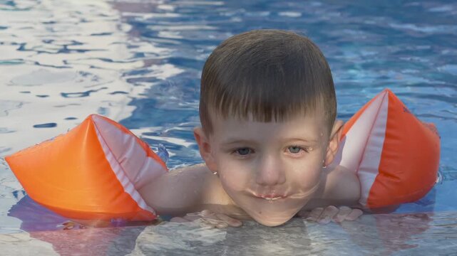 Happy smiling child in orange inflatable arm bands smiles at camera beside clear blue swimming pool. Learning to swim, family vacation and positive childhood memories concept. Smiling boy 