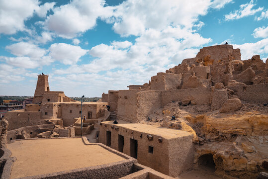 View of Shali Fortress, an ancient mud-brick structure with textured walls and towers under a bright blue sky with scattered clouds in Siwa, Marsa Matrouh Governo, Egypt.
