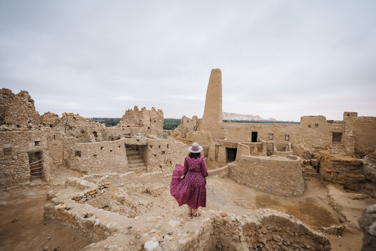 View of a woman in a purple dress and hat standing amidst the ancient mud-brick ruins of Shali Fortress under a cloudy sky in Siwa, Marsa Matrouh Governo, Egypt.