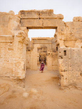 View of a woman in a pink dress walking through the ancient stone ruins of the Amon Temple with weathered limestone blocks and sandy ground in Siwa, Marsa Matrouh Governo, Egypt.