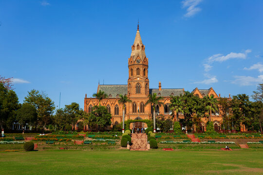 View of Government College University Lahore with its historic red brick gothic architecture, central clock tower, lush green lawns, and palm trees in Lahore, Punjab, Pakistan.