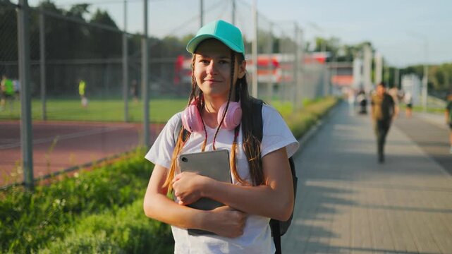 Smiling Zalpha teenager girl holding digital tablet near outdoor sports field.