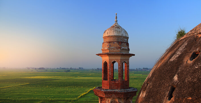 View of the historic brick minaret of the Shrine of Abdul Nabi surrounded by lush green fields under a clear sky at sunset in Kotli Maqbara, Punjab, Pakistan.
