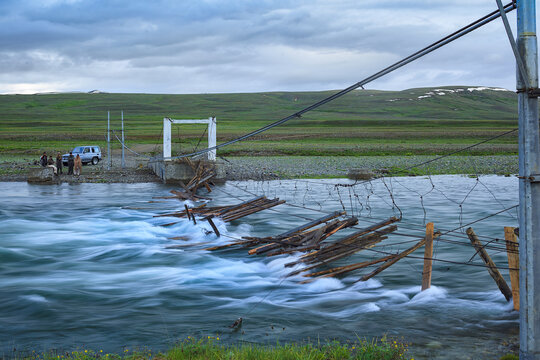 View of the abandoned bridge at Bara Pani in Deosai National Park with rushing river water, green hills, and people standing near a vehicle in Skardu, Gilgit Baltistan, Pakistan.