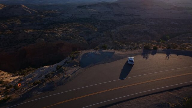 Aerial drone footage Utah Scenic Byway 12 captures white SUV parked at scenic pullout with person standing at canyon edge enjoying vast red rock landscape during golden hour sunset light.