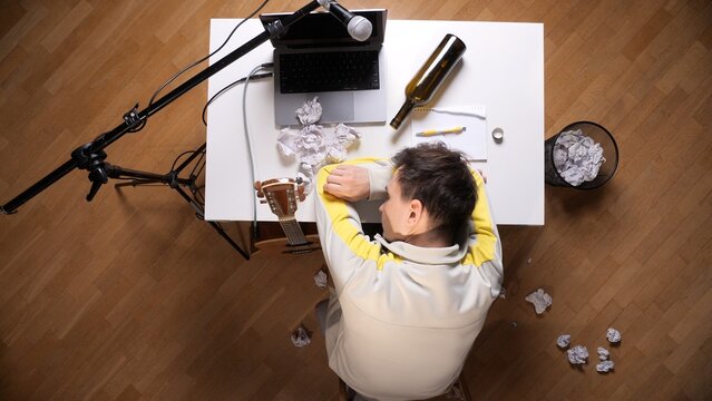 Musician lying on a white desk, head on arms, surrounded by an empty wine bottle, crumpled papers, a laptop with a microphone, and an acoustic guitar, signifying creative block and exhaustion