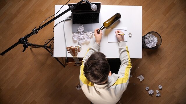 Young man experiencing writer's block at his desk, surrounded by crumpled papers and an empty wine bottle, symbolizing frustration and lack of inspiration while attempting to write music