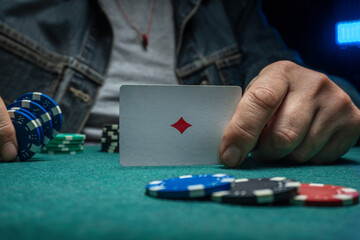 A person holds a playing card at a poker table. Various chips are stacked on the green tabletop. The setting is inside a casino at night with dim lighting © Віталій Б.