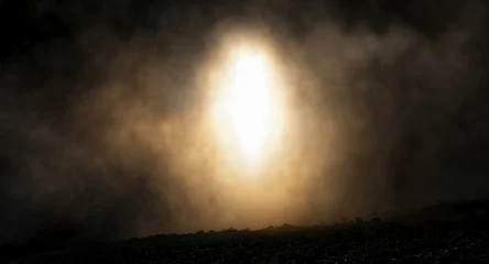 A bright, glowing oval light shines through dense fog and smoke over a dark, rocky ground, creating a dramatic, ethereal atmosphere. © ginettigino