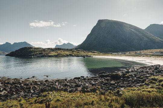 View of Hovden beach with rocky shoreline, calm sea, and dramatic mountains under a clear sky in Hovden, Nordland, Norway.