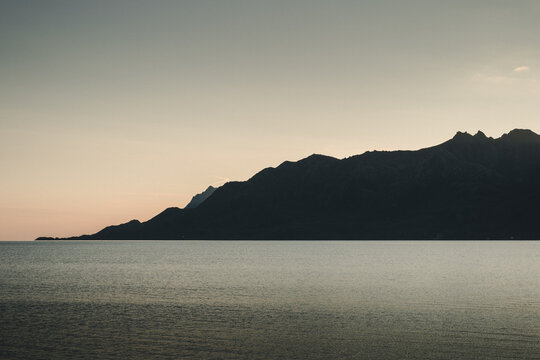 Holmstad, Nordland, Norway - 30 August 2025: View of the silhouette of mountains against a soft sunset sky over the calm sea.