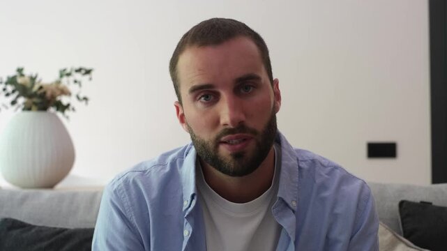 Young bearded man sitting on a couch and having a serious conversation while looking at the camera, showing various emotions from concern to sadness and then actively explaining something