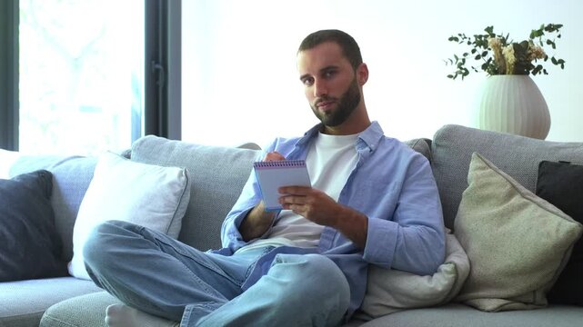 Focused young man sitting cross-legged on a comfortable couch at home, writing down thoughts, plans, or a to-do list in a small notebook and then looking at the camera with a smile