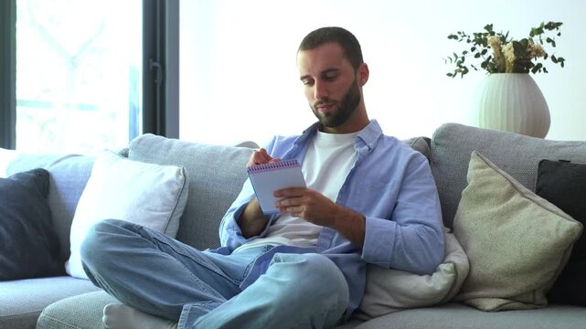 Thoughtful young man sitting comfortably on a sofa in his modern living room, writing down ideas and plans in a small spiral notebook, then looking thoughtfully towards the camera
